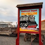 Ritzville WA 21 A colorful sign for the Railroad Depot Museum and Antique Farm Equipment stands in front of vintage farm machinery and a gravel lot under a cloudy Ritzville, WA sky.