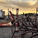 Ritzville WA 22 Rusty vintage farm equipment sits on gravel under a dramatic sunset sky in Ritzville, WA, with a small town and silo buildings visible in the background. The scene exudes an antique, nostalgic atmosphere.