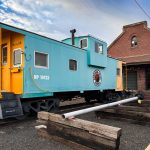 Ritzville WA 24 A vintage turquoise and yellow caboose labeled NP 10425 is displayed on tracks beside Ritzville’s historic brick train station in WA, under a partly cloudy sky. Wooden beams and a metal pole are in the foreground.