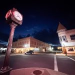 Ritzville WA 8 A street clock shows 9:00 PM on a quiet downtown Ritzville, WA intersection at night, with historic brick buildings, glowing shop lights, and a car driving by under a deep blue sky.