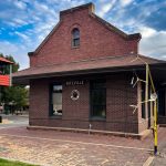 Ritzville WA 29 A historic red-brick train station in Ritzville, WA, labeled RITZVILLE, features a decorative round window and a small railroad crossing signal nearby under a partly cloudy sky.