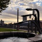 Ritzville WA 27 A metal fountain with arrow-shaped spouts pours water into a circular stone basin at sunset in Ritzville, WA. Industrial buildings, a lamp post, and railroad tracks appear in the background under a partly cloudy sky.