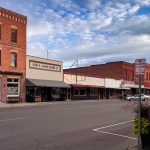 Ritzville WA 18 A quiet small-town street in Ritzville, WA, features historic brick buildings, storefronts, and angled parking. A few cars are parked under a partly cloudy blue sky, with a "no parking" sign and a planter in the foreground.