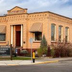 Ritzville WA 15 A historic brick building labeled Carnegie Library stands on a street corner in Ritzville, WA, with striped awnings, large windows, and a statue out front. A black sign and landscaped bushes complete the charming scene.