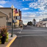 Ritzville WA 12 A quiet Ritzville, WA small-town street with a “Ritz” theater sign, American flags, a lamppost, and potted plants on the corner under a blue sky. The empty street is lined with charming buildings on both sides.