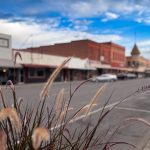 Ritzville WA 11 Tall grass in focus in the foreground with an empty small-town street, historic brick buildings, parked cars, and a partly cloudy blue sky in the background of Ritzville, WA.