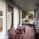 Ritzville WA 33 A wooden porch with red Adirondack chairs and small tables is attached to a white house with decorative trim in Ritzville, WA. Plaques hang on the wall, and a motorcycle is visible in the background near a fence and trees.
