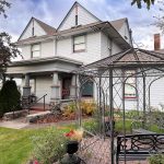 Ritzville WA 34 A large white two-story house in Ritzville, WA, features a covered front porch, lush landscaping, and a green lawn. In the foreground, a metal gazebo with a bench and decorative accents offers a charming outdoor retreat.