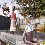 Ritzville WA 36 Two whimsical metal sculptures sit in a cart in Ritzville, WA; one figure in front has wide eyes and a blue shirt, while a second figure at the back holds flowers. A house and tree are visible in the blurred background.