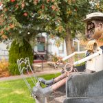 Ritzville WA 37 A metal sculpture of a person with a mustache and hat sits in a black car-shaped structure outdoors in Ritzville, WA, with a tree and houses visible in the background.