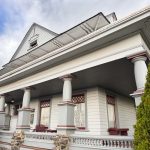 Ritzville WA 35 A large, white, two-story house in Ritzville, WA, features a wide porch with thick columns, detailed red trim, stone accents, and surrounding shrubs under a cloudy sky.