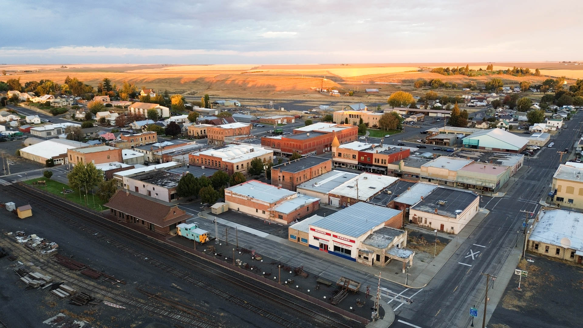 Aerial view of Ritzville, WA, with brick buildings, streets, and railroad tracks in the foreground; open fields and farmland stretch out in the background under a partly cloudy sky.
