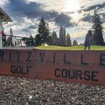 Ritzville WA 39 Wooden sign reading Ritzville Golf Course in the foreground, with a golfer swinging a club on green grass in the background, trees, and a partly cloudy WA sky with sunlight.