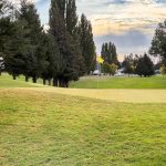 Ritzville WA 41 A golf course green in Ritzville, WA, with a yellow flagstick, is surrounded by neatly mowed grass and tall pine trees, under a partly cloudy sky with sunlight breaking through.