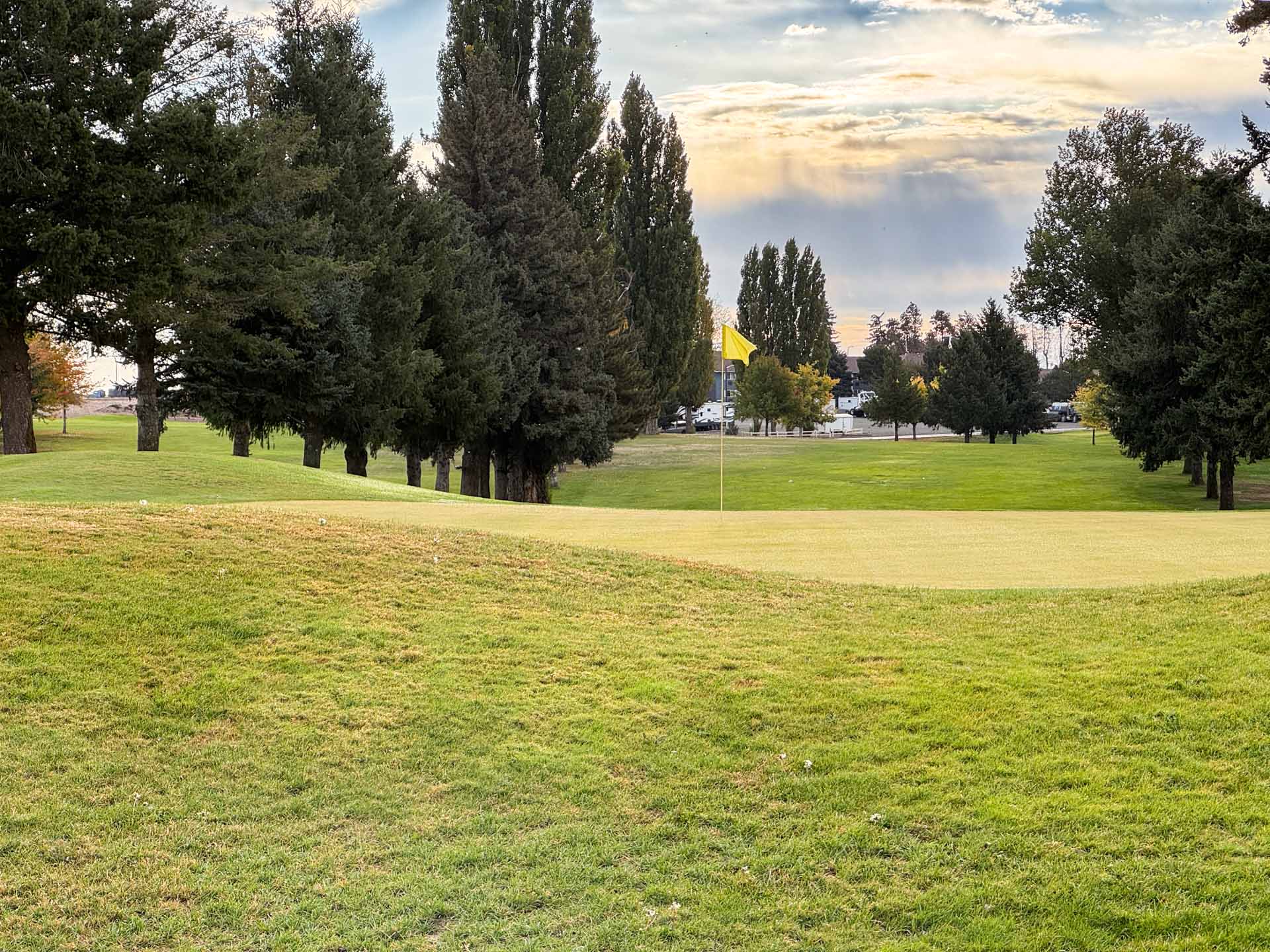 A golf course green in Ritzville, WA, with a yellow flagstick, is surrounded by neatly mowed grass and tall pine trees, under a partly cloudy sky with sunlight breaking through.