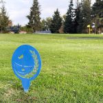 Ritzville WA 40 A blue metal golf tee marker with a flag and laurel design sits on green grass at Ritzville Golf Course, WA, with trees in the background and a yellow flag visible in the distance.