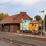 Ritzville WA 20 A historic red brick train station in Ritzville, WA stands beside railroad tracks, with a turquoise and yellow caboose displayed outside, surrounded by a fence and outdoor seating under a cloudy sky.